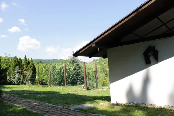 Ugao zgrade hotela s ciglenom stazom, uz nju ograđeni ispust s mladim crnogoricama i brežuljci u daljini.